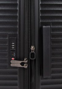 Gros plan sur une valise rigide noire avec double zip, verrou à combinaison TSA et une poignée latérale sur une surface nervurée.