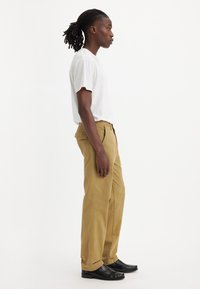 Pantaloni drepți bej, din bumbac, asociați cu un tricou alb cu mânecă scurtă. Pantofi negri completează ținuta. Vedere din profil lateral.