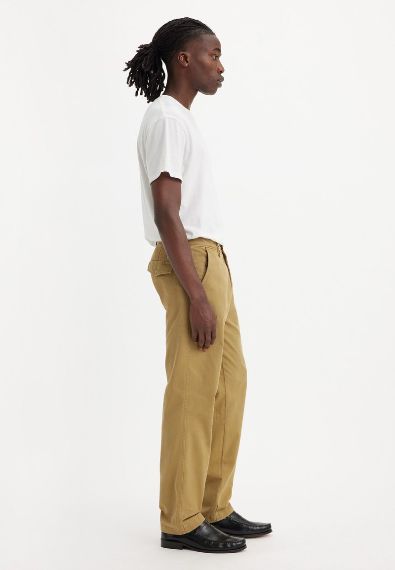 Pantalones rectos de color beige hechos de algodón, combinados con una camiseta blanca de manga corta. Zapatos negros completan el look. Vista de perfil lateral.