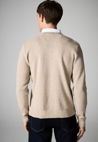 Pulover bej din tricot, cu un finisaj texturat, mâneci și tiv ribbate, guler tip crew și o croială lejeră, purtat cu jeans negri.