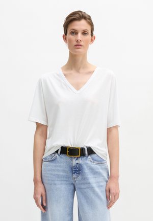 Femeie purtând un tricou simplu alb cu gât în formă de V, băgat într-o pereche de blugi albastru deschis, cu o curea neagră ce are o cataramă mare pătrată aurie.