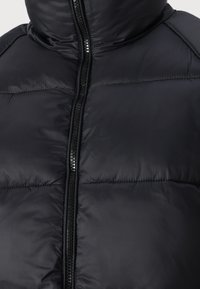 Casaco puffer preto com gola alta, fecho de correr e textura acolchoada. O tecido parece suave e brilhante, com detalhes mínimos.