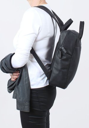 Zaino nero in pelle con forma rettangolare, tasca frontale con zip e spallacci regolabili, indossato su una spalla sopra una camicia bianca.