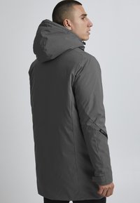 Parka longue grise avec capuche, en matériau lisse et résistant à l'eau. Comprend des poches latérales et un design effilé et épuré.