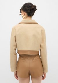 Beige cropped frakke med en tekstureret ulden overdel, brun kunstlæder-bælte, og tætsiddende brune shorts med baglommer.