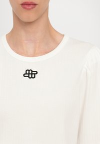 Hvit ribbestrikket topp med rund hals, med et svart brodert design i midten. Myk tekstur og puffermer er synlige.