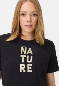 Sort T-shirt lavet af blød bomuld med rund hals, med en vertikal, metallisk gul tekstgrafik, der læser "EMPOWERED BY NATURE."