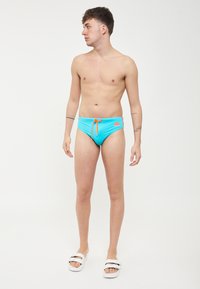 Bañador azul con cordón naranja y detalle de logo, diseño ajustado, textura suave, combinado con sandalias blancas.