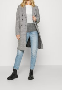 Grauer und schwarz karierten Wollmantel, doppelreihig mit großen Knöpfen. Trägt man über grauem Pullover und blauen Jeans, kombiniert mit schwarzen Stiefeletten.