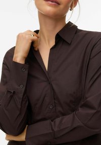 Chemise marron foncé à boutons, col structuré, coupe ajustée et poignets boutonnés, avec une texture de tissu lisse et sans motifs visibles.