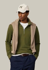 Sudadera verde de manga larga con cremallera, con un suéter de punto beige sobre los hombros, combinado con pantalones azul marino y una gorra de béisbol color crema.