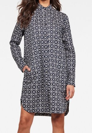 Femme portant une robe à motifs géométriques noire et blanche, longueur genou, boutonnée, avec des manches longues et une main dans la poche.