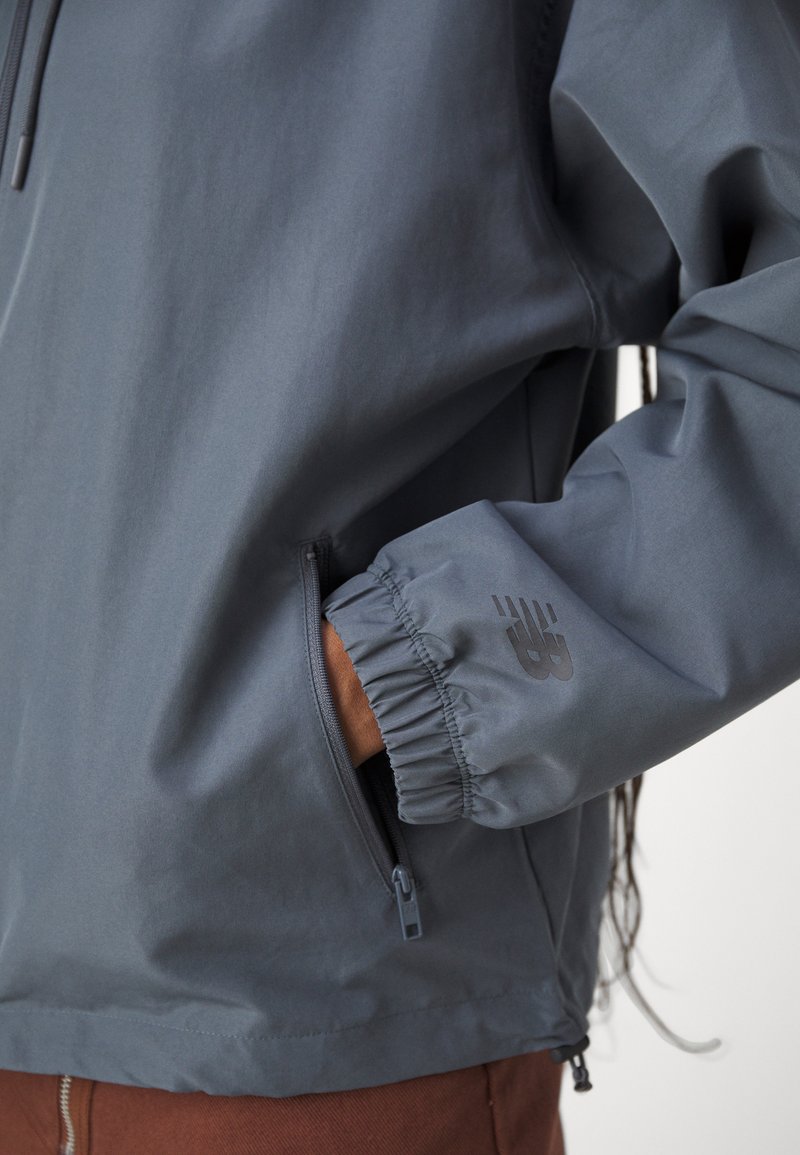 Person, der eine dunkelgraue Jacke trägt, mit einer Hand in der Reißverschlusstasche, sichtbarem elastischen Bündchen und einem Logo auf dem Ärmel über braunen Hosen.
