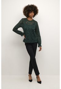 Top in pizzo a maniche lunghe di colore verde scuro con motivi floreali, abbinato a pantaloni neri su misura e décolleté neri.