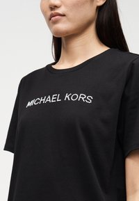 T-shirt preta de algodão com gola redonda e mangas curtas, adornada com cristais brancos que formam a palavra "MICHAEL KORS" ao longo do peito.