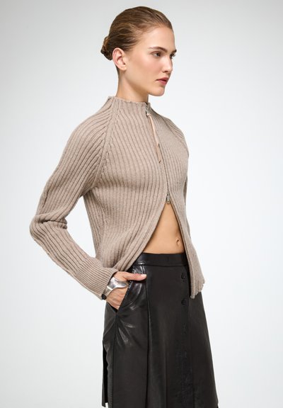 Beige csíkozott zipzáras pulóver magas gallérral, hosszú ujjakra és nyitott elülső részre tervezve. Fekete bőr szoknyával párosítva.