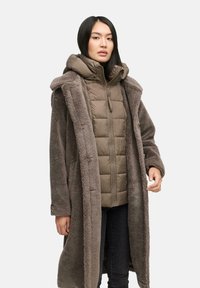 Doudoune marron atténué avec un design matelassé, portée sous un long manteau doux en fourrure ressemblante de teinte assortie avec des boutons.