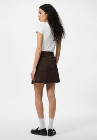 Jupe mini marron avec une taille cintrée et fermeture éclair, associée à une chemise blanche à manches courtes et des chaussures noires avec des chaussettes blanches.