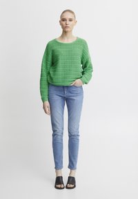 Grüner Strickpullover mit strukturiertem Muster, getragen zu hellblauen Skinny-Jeans und schwarzen Slide-Sandalen. Weißer Hintergrund.
