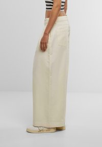 Wide-leg trousers in soft beige cotton, featuring a back pocket and a smooth texture, paired with white sneakers displaying handwritten details.
