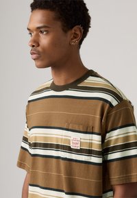 Camiseta de rayas marrones con detalles en blanco y verde. Presenta un bolsillo en el pecho izquierdo con una marca en rojo. Está confeccionada con un material de algodón suave.