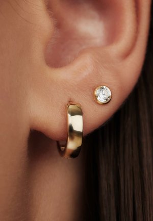 Boucle d'oreille en or et une petite puce ronde avec une pierre claire, présentées contre un gros plan de peau et de cheveux. Surface lisse, finition brillante.