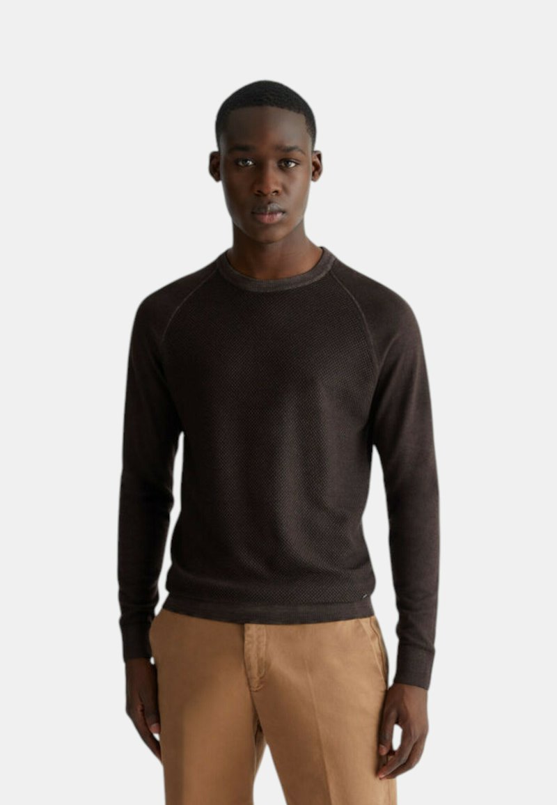 Maglione a collo alto castano scuro con un motivo testurizzato, scollatura aderente e maniche raglan, indossato sopra pantaloni beige chiaro.