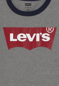 Camiseta gris con cuello marino que presenta un gran logotipo gráfico rojo en forma de alas de murciélago, donde se muestra "Levi's" en letras blancas en negrita.