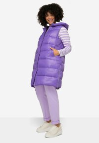 Gilet rembourré violet avec capuche et poches zippées, superposé sur une chemise à rayures à manches longues, associé à un pantalon violet clair et des baskets beiges.