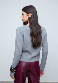 Vrouw met lang zwart haar in een grijze trui en bordeaux leren broek, met een zwarte bloemenoorbel en gestudde zwarte armbanden, die naar links kijkt.
