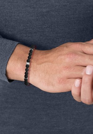 Bracciale in perle nere caratterizzato da perline matte lisce e accenti metallici. Il bracciale è indossato su un polso contro uno sfondo di maglione scuro.