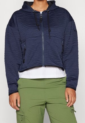 Persoon draagt een marineblauwe, gestructureerde hoodie met rits en trekkoorden over een wit overhemd en groene cargobroek.