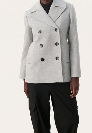 Personne portant un manteau en laine gris clair à double boutonnage avec de gros boutons noirs et un pantalon cargo noir avec des poches, debout.
