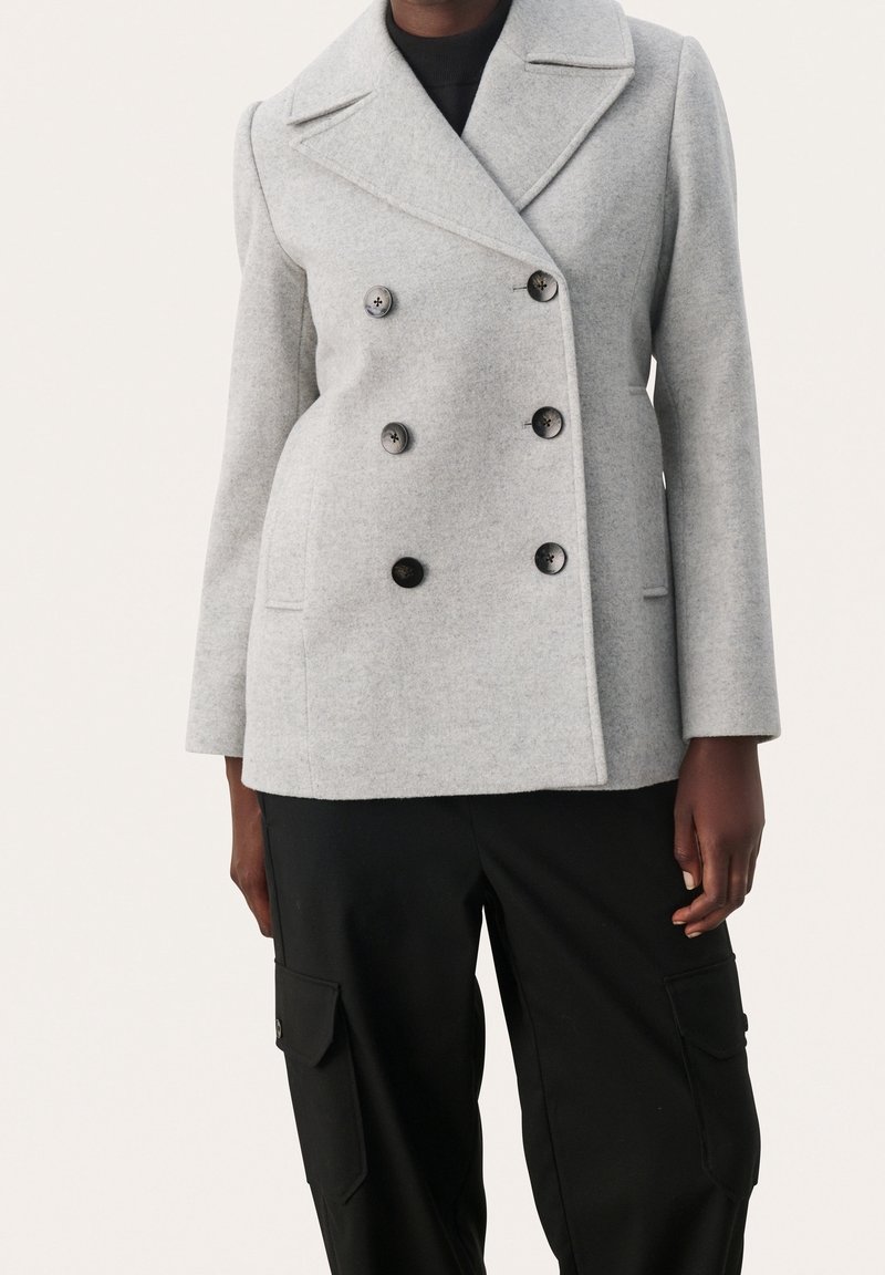Personne portant un manteau en laine gris clair à double boutonnage avec de gros boutons noirs et un pantalon cargo noir avec des poches, debout.