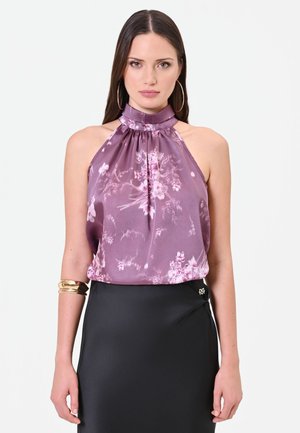 Frau trägt ärmelloses lila florales Neckholder-Top mit hohem Kragen und schwarzem Rock, ergänzt durch große Creolen und goldene Armreifen.