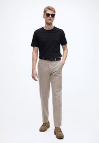 Svart t-shirt med kort ärm, beige texturerade byxor och bruna slip-in skor. Modellen bär mörka solglasögon och står mot en vit bakgrund.