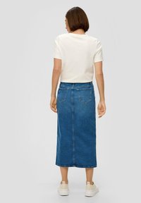 Blauwe denim midi-rok met een rechte snit, twee achterzakken en een zoom die tot halverwege de kuit reikt, gedragen met een wit shirt en lichte sneakers.