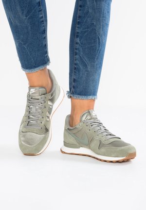 Pieds portant des baskets Nike vert clair assorties à un jean bleu court sur fond blanc.