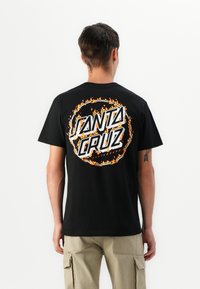 Sort bomulds t-shirt med et cirkulært grafik, der viser "Santa Cruz" i hvidt og orange flammer, sat mod en ensfarvet baggrund.