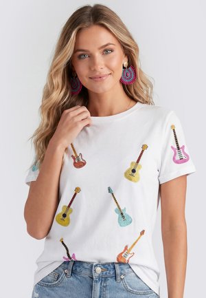 Vrouw met lang blond haar die een wit T-shirt draagt versierd met kleurrijk geborduurde gitaren en een blauwe spijkerbroek, zacht glimlachend.