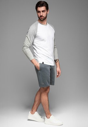 Chemise à manches longues blanche et grise associée à un short gris. Le short possède des poches et une étiquette de logo. Modèle portant des baskets blanches.