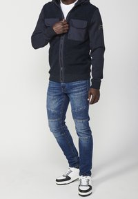Marinblå zip-up hoodie med texturerade, furriga paneler och två bröstfickor. Kombinerad med smal passform blå jeans och svart-vita sneakers.