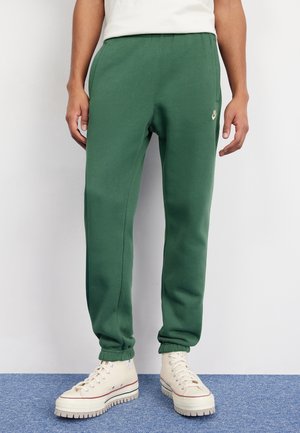 Mann trägt grüne Nike Jogginghose und weiße High-Top-Sneaker, steht auf blauem Teppich vor weißer Wand