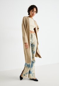 Beige cardigan, kort beige topp, ljusblåtie-dye vida jeans med sidoflikar, och svarta spetsiga flats. Slät textur, avslappnad passform.