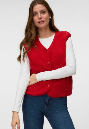 Kvinde iført en lys rød fleecevest over en hvid langærmet skjorte og blå jeans, der står mod en ensfarvet lys baggrund.
