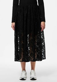 Jupe maxi en dentelle noire avec un motif floral, taille élastique, design superposé et texture transparente, associée à des baskets argentées.