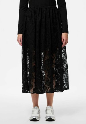 Gonna maxi in pizzo nero con motivo floreale, vita elastica, design a strati e texture trasparente, abbinata a sneakers argento.