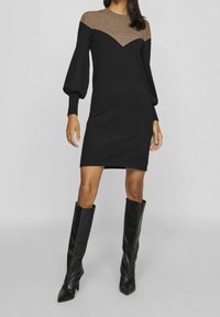 Robe noire tricotée avec un empiècement triangulaire marron, des manches bouffantes et une silhouette ajustée. Portée avec des bottes en cuir noires montantes.