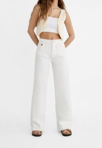 Kobieta w białych, szerokich spodniach o wysokim stanie, z przednimi kieszeniami, białym topie crop, beżowej kamizelce zapinanej na guziki i beżowych sandałach.