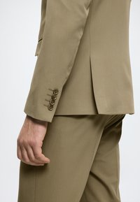 Lichtbruin colbert met een getailleerde pasvorm, vier knopen op de mouwen en een split aan de achterkant, gemaakt van een gladde, gestructureerde stof.